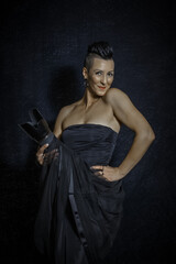 A beautiful woman with a mohawk with black short hair in a black top without straps and a black skirt holds a camera with a long lens and smiles coquettishly