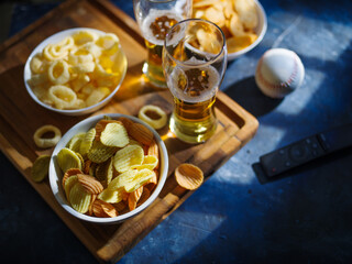 On a wooden tray are potato chips, onion rings and two glasses of light beer. TV remote and golf ball. High angle view. Relaxation, watching sports games on TV with friends.