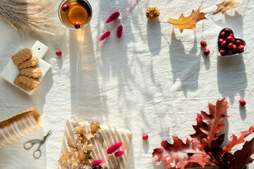 Autumn decorations - cranberry, cookies. tea and dry oak leaves. Flat lay, top view on uncolored textile. Fall background with copy-space. Sunlight, long shadows. Wrapped gift with bunny tail grass.