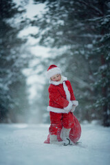 Little funny child dressed in Santa Claus red costume bringing presents in winter snowy forest. Christmas Eve.