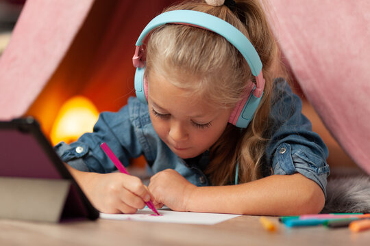 Child girl draws with felt-tip pens in headphones with a tablet. Online lesson for preschoolers.