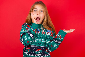 Positive glad little kid girl wearing knitted sweater christmas over red background says: wow how exciting it is, indicates something.  One hand on his head and pointing with other hand.