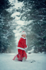 Little funny child dressed in Santa Claus red costume bringing presents in winter snowy forest. Christmas Eve.