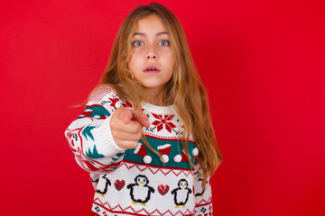 Shocked little kid girl wearing knitted sweater christmas over red background points at you with stunned expression