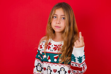 little kid girl wearing knitted sweater christmas over red background shows middle finger bad sign asks not to bother. Provocation and rude attitude.