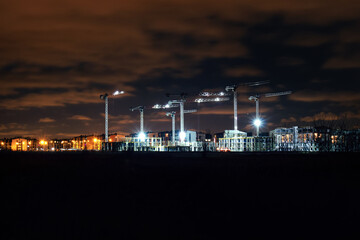 Tower cranes on construction of buildings at night, illuminated by bright lanterns, view from afar