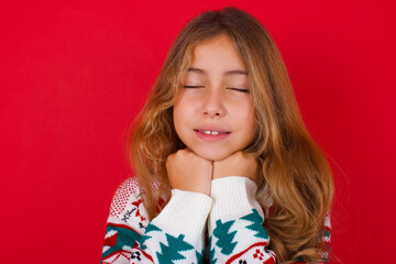 Cheerful little kid girl wearing knitted sweater christmas over red background has shy satisfied expression, smiles broadly, shows white teeth, People emotions