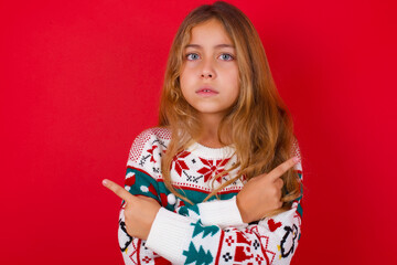Obraz na płótnie Canvas Serious little kid girl wearing knitted sweater christmas over red background crosses hands and points at different sides hesitates between two items. Hard decision concept