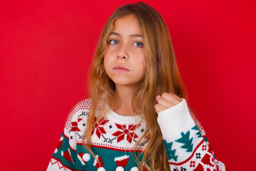 little kid girl wearing knitted sweater christmas over red background shows fist has annoyed face expression going to revenge or threaten someone makes serious look. I will show you who is boss
