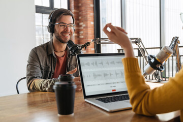 Radio podcast concept. Cheerful male radio guest looking at female host, wearing headphones, answering a question for audio podcast in studio. Woman interviewing famous influencer for channel content