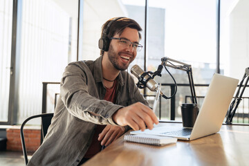 Young man host in headphones and glasses enjoying podcasting in studio, speaking into a microphone, holding a pen, using laptop. Handsome podcaster laughing while streaming live audio podcast - obrazy, fototapety, plakaty