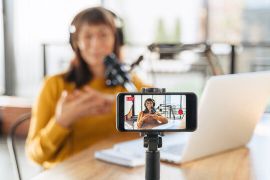 Female vlogger live streaming podcast using microphone, laptop and cellphone on tripod. Selective focus on smartphone camera screen with woman podcaster recording and broadcasting live video. Close-up