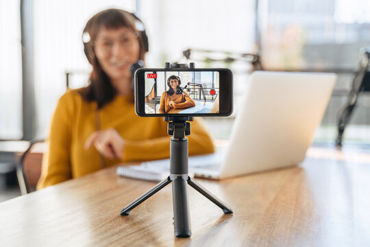 Woman radio host in headphones streaming live video podcast using microphone and laptop in studio. Female youtuber looking at camera, shooting vlog. Selective focus on smartphone camera. Close-up