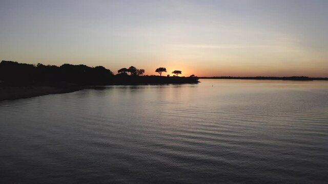 The drone flies toward and over a peninsula with the sun setting the distance.  The drone starts out low over the Texas lake.  Eventually the camera move reveals a boat on the water.