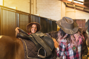 Two cowgirl women preparing to ride a horse in a stable, southern usa hats and jeans, vertical photo