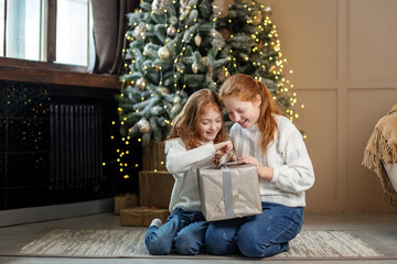 Two little sisters open gift boxes under the tree in the room. Winter holidays concept, merry christmas and family