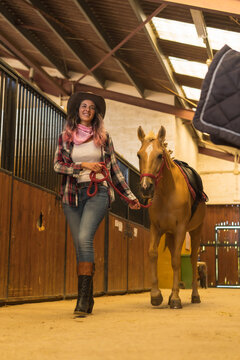 Caucasian cowgirl woman posing and smiling with a horse in a stable, southern usa hats, pink plaid shirt and jeans