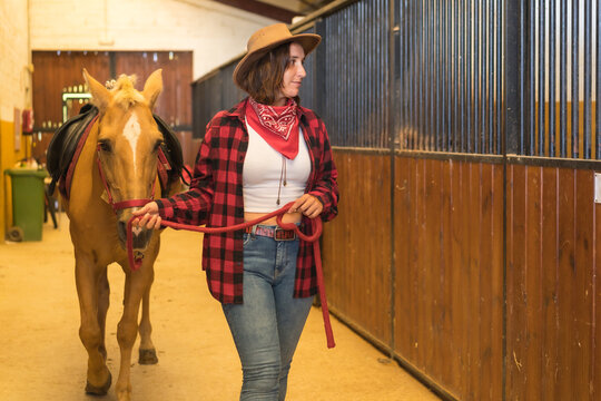 Caucasian brunette cowgirl woman strolling with a horse in a stable, wearing American cowboy hat, red plaid shirt and jeans