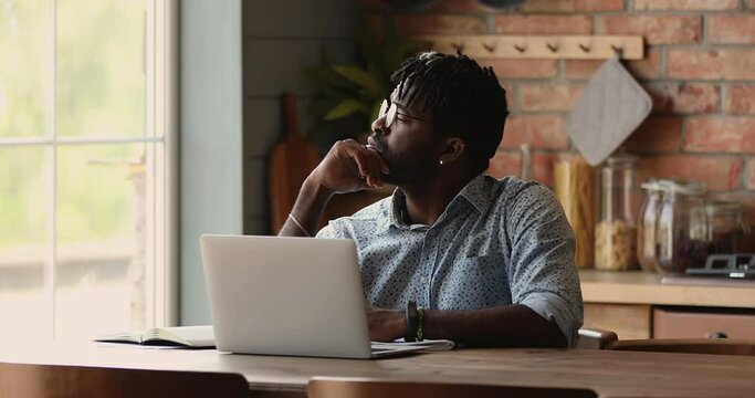 Thoughtful African man in glasses dreadlocks hairstyle sit at table writing making notes in copybook studying thinking learning on-line use laptop. Webinar, self-education using modern tech concept
