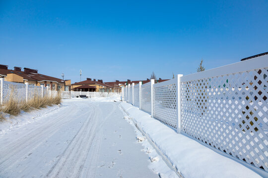 Snowy Road In A Cottage Village. White Plastic Fence In A Modern Cottage Village On A Clear Winter Day. Snow Drifts In Front Of A Vinyl Fence Against A Blue Sky