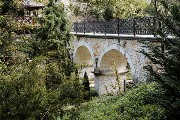 Stone bridge with ornate black fence. Attraction of the area. Bridge in the spruce garden. Walk in the fresh air. Luxurious bridge made of white stone.