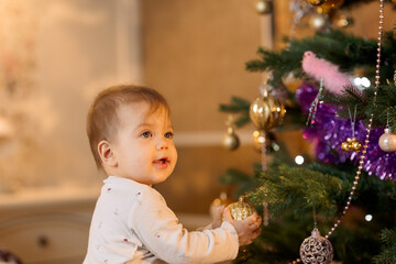 Fototapeta na wymiar child in white clothes at home next to a christmas tree holds a christmas ball and smiles 