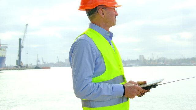 Sea industry port ship safety. Smiling general manager in vest and helmet with tablet and portable radio looks at new vessel in dockyard