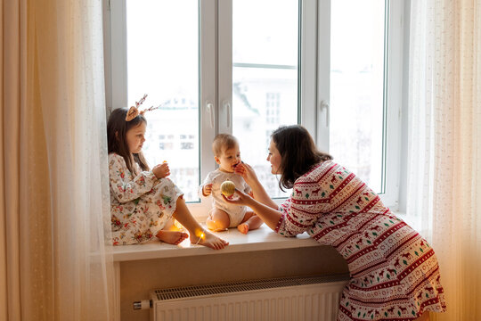 Family Christmas photo. Mom and two daughters are playing on the windowsill. It's winter outside and snow is falling