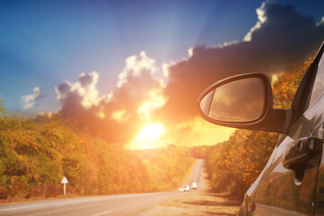 Close-up of a car mirror with a blurred countryside road and a sky with a sunset