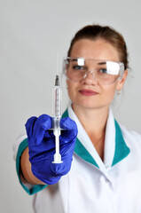 Female doctor with protective glasses and gloves holding a syringe against a grey background