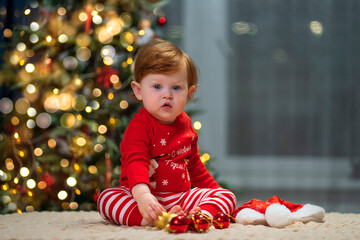 A baby with red hair in a red Christmas costume helps to decorate the Christmas tree. The inscription on the clothes - Happy New Year.