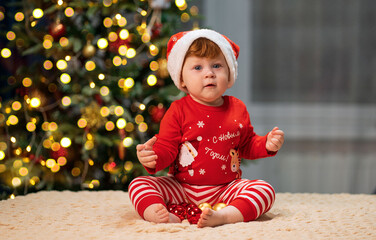 A small child in a red suit and a santa claus hat against the background of a Christmas tree. Inscription on clothes: happy new year