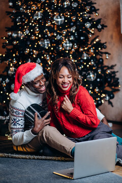happy african american couple congratulating and making video calls on laptop, sitting on the floor with a gift against the background of a christmas tree, new technologies vertical photo