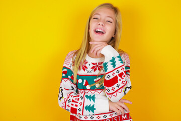 Optimistic caucasian little kid girl wearing knitted sweater christmas over yellow background keeps hands partly crossed and hand under chin, looks at camera with pleasure. Happy emotions concept.