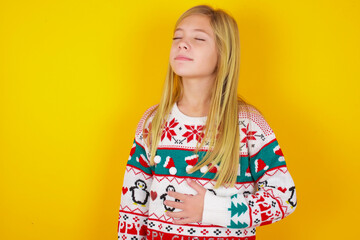caucasian little kid girl wearing knitted sweater christmas over yellow background touches tummy,...