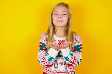 caucasian little kid girl wearing knitted sweater christmas over yellow background holding something with open palms, offering to the camera.