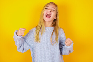 caucasian kid girl wearing blue knitted sweater over yellow background celebrating surprised and...