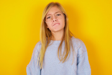 caucasian kid girl wearing blue knitted sweater over yellow background making grimace and crazy...