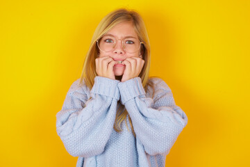 Fearful caucasian kid girl wearing blue knitted sweater over yellow background  keeps hands near...