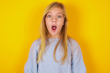 Oh my God. Surprised caucasian kid girl wearing blue knitted sweater over yellow background  stares...