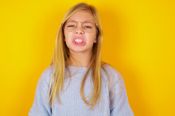 caucasian kid girl wearing blue knitted sweater over yellow background  keeps teeth clenched, frowns face in dissatisfaction, irritated because of much duties.