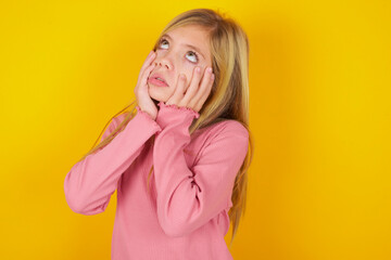 caucasian little kid girl wearing long sleeve shirt over yellow background keeps hands on cheeks...