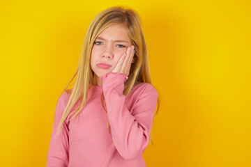 Sad lonely caucasian little kid girl wearing long sleeve shirt over yellow background touches cheek with hand bites lower lip and gazes with displeasure. Bad emotions