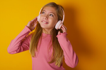 Joyful caucasian little kid girl wearing long sleeve shirt over yellow background sings song keeps hand near mouth as if microphone listens favorite playlist via headphones