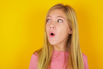 Shocked caucasian little kid girl wearing long sleeve shirt over yellow background look empty space...