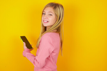 Rear view photo portrait of caucasian little kid girl wearing long sleeve shirt over yellow background using smartphone smiling