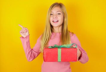 Cute little blonde girl standing over yellow background holding a present and pointing aside at empty copyspace.