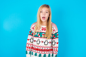 little kid girl wearing knitted sweater christmas over blue background having stunned and shocked look, with mouth open and jaw dropped exclaiming: Wow, I can't believe this. Surprise and shock