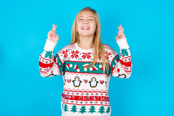 Joyful little kid girl wearing knitted sweater christmas over blue background clenches teeth, raises fingers crossed, makes desirable wish, waits for good news, I have to win.