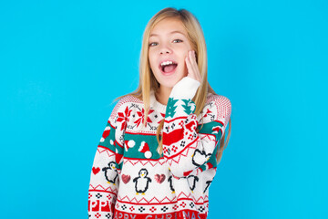 Shocked, astonished caucasian little kid girl wearing knitted sweater christmas over blue background looking surprised in full disbelief wide open mouth with hand near face. Positive emotion 